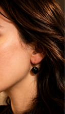 Black onyx checker cut heart drop earrings with gold hook closures laid flat on a light grey surface, showing the faceted stone detail and gold bezel setting.